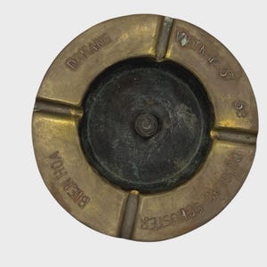 May include: A vintage brass ashtray with a circular design. The ashtray has four grooves for resting cigarettes and features the engraved text "DANANG", "BIEN HOA", "VIETNAM 67", and "DOUGLAS SCHUSTER 68". The center is a darker, aged color.