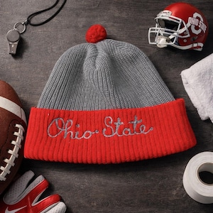 May include: A gray and red knit beanie with "Ohio State" embroidered in silver. The hat has a red pom-pom on top. A red football helmet and a football are also in the image, suggesting a sports theme.