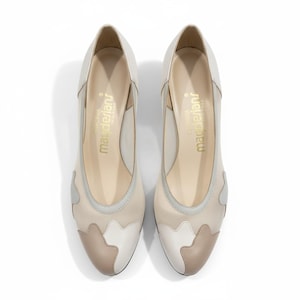 May include: A pair of cream-colored women's pumps with a unique design. The shoes feature a combination of beige, white, and gray accents. The brand name "magderians" is printed in gold on the insole. The heels are a moderate height.