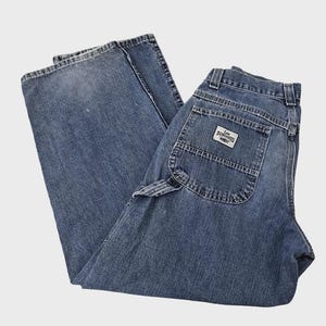May include: Blue denim Lee Dungarees work pants. The pants have a classic five-pocket design, a hammer loop, and a back pocket with a Lee Dungarees label. The denim has a faded appearance.