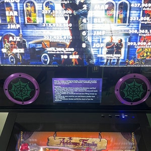 May include: A pinball machine with a Addams Family theme. The machine has a black cabinet with a purple and green spiderweb design on the speakers. The backglass features the Addams Family characters and a high score list. The playfield is visible with a lit candle and a sign that reads "Addams Family".