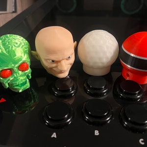 May include: Four different joystick toppers, including a green skull with red eyes, a bald man with a sinister smile, a white golf ball, and a red and black striped sphere.