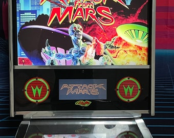May include: A vintage pinball machine with a glass top and a silver metal frame. The backglass features a colorful image of a spaceship and aliens with the text "Attack from Mars" and "Bally". The playfield is visible below the backglass and features a green and red "W" logo.