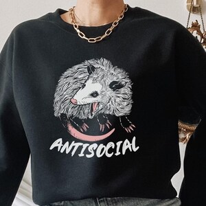 K&ouml;nnte beinhalten: Schwarzer Pullover mit einem Aufdruck eines Opossums mit dem Text "ANTISOCIAL". Das Model tr&auml;gt eine blaue Jeans mit Rissen.