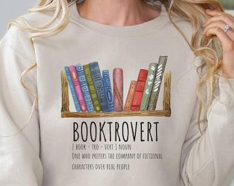May include: A beige sweatshirt with a graphic design of a bookshelf with books and the text "BOOKTROVERT [BOOK - TRO - VERT] NOUN ONE WHO PREFERS THE COMPANY OF FICTIONAL CHARACTERS OVER REAL PEOPLE"