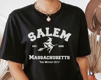 May include: Black t-shirt with a white graphic of a witch flying on a broomstick. The graphic includes the text "Salem, Est. 1626, Massachusetts, 'The Witchy City'".