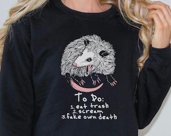 Könnte beinhalten: Schwarzes Sweatshirt mit einer weißen und rosa Illustration eines Opossums mit dem Text "To Do: 1. eat trash 2. scream 3. fake own death"