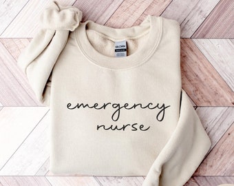 May include: A sand-colored sweatshirt with the text "emergency nurse" written in black cursive script.