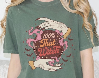 May include: A moss green t-shirt with a graphic of a crystal ball with two hands holding it. The crystal ball has stars and bats around it and the text "100% That Witch".