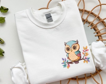 Könnte beinhalten: Weißes T-Shirt mit Rundhalsausschnitt und einem bunten Cartoon-Eulen-Motiv, das auf einem Ast mit Blättern sitzt. Die Eule hat blaue Augen und einen braunen Körper mit blauen und grünen Federn.