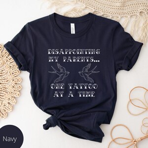 May include: Navy blue t-shirt with white text that reads "Disappointing my parents... One tattoo at a time". The text is in a vintage style font and is accompanied by two stylized bird tattoos.