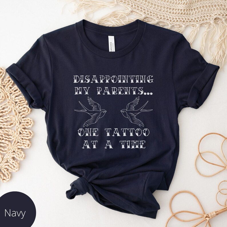 May include: Navy blue t-shirt with white text that reads "Disappointing my parents... One tattoo at a time". The text is in a vintage style font and is accompanied by two stylized bird tattoos.