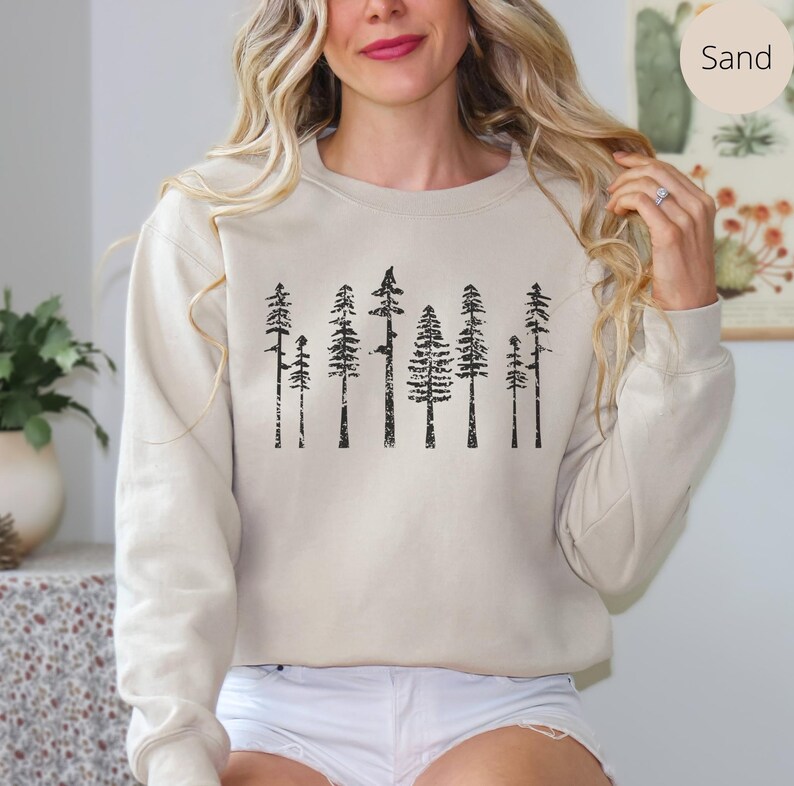 May include: A beige sweatshirt with a black graphic print of a row of trees. The sweatshirt has a crew neck and long sleeves. The text "Sand" is visible in the upper right corner of the image.