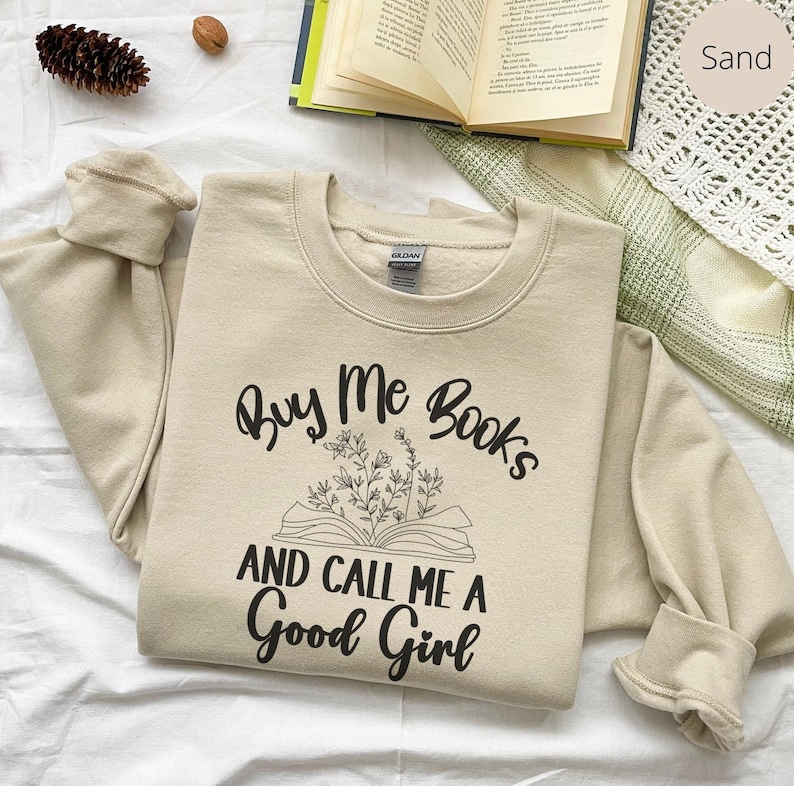 May include: A sand-colored sweatshirt with the text "Buy Me Books and Call Me a Good Girl" printed in black. The text is accompanied by a black line drawing of an open book with flowers.