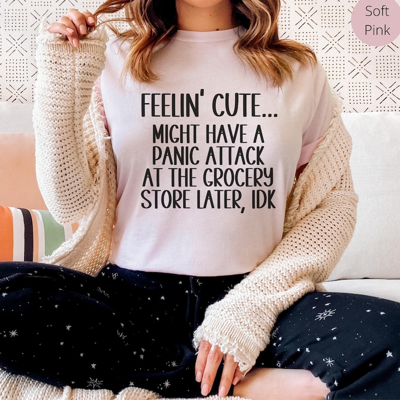 May include: A soft pink t-shirt with the text "FEELIN' CUTE... MIGHT HAVE A PANIC ATTACK AT THE GROCERY STORE LATER, IDK" printed in black.