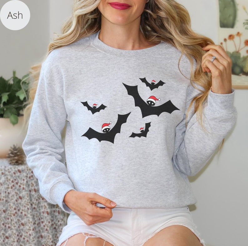 K&ouml;nnte beinhalten: Ein graues Sweatshirt mit einem weihnachtlichen Design mit schwarzen Flederm&auml;usen, die Weihnachtsm&uuml;tzen tragen. Die Flederm&auml;use fliegen in verschiedene Richtungen.