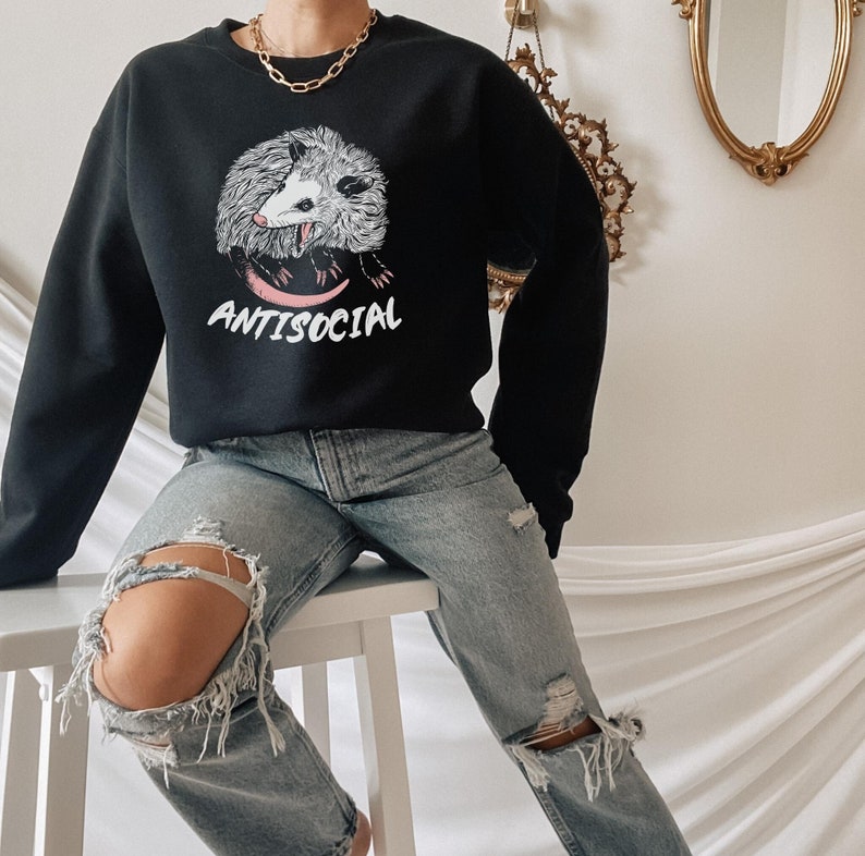 K&ouml;nnte beinhalten: Schwarzer Pullover mit einem Aufdruck eines Opossums mit dem Text "ANTISOCIAL". Das Model tr&auml;gt eine blaue Jeans mit Rissen.