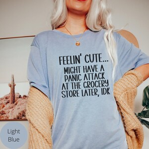 May include: Light blue t-shirt with the text "Feelin' Cute... Might have a panic attack at the grocery store later, idk" printed in black.