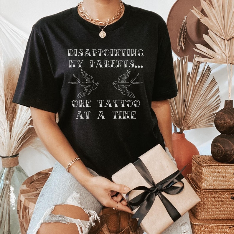 May include: Black t-shirt with the text "DISAPPOINTING MY PARENTS... ONE TATTOO AT A TIME" and two stylized birds.