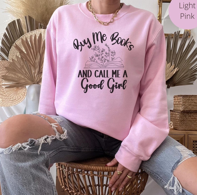 May include: A light pink sweatshirt with the text "Buy Me Books And Call Me A Good Girl" printed on the front. The text is in black and features a stylized illustration of a book with flowers.