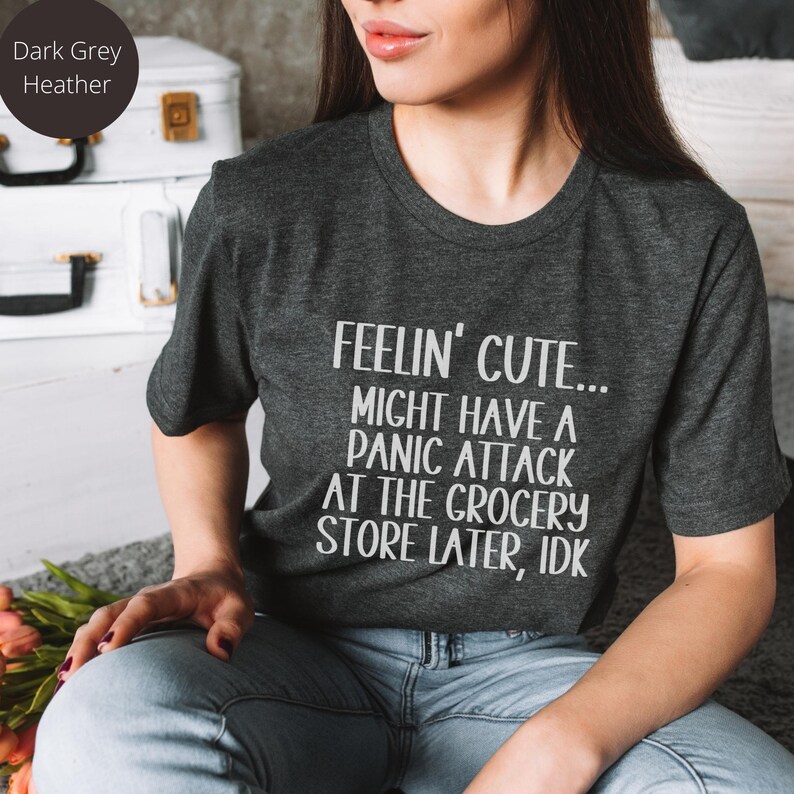 May include: A dark heather grey t-shirt with the text "FEELIN' CUTE... MIGHT HAVE A PANIC ATTACK AT THE GROCERY STORE LATER, IDK" printed in white.