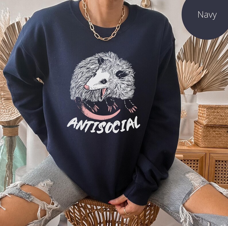 K&ouml;nnte beinhalten: Ein marineblauer Pullover mit einem wei&szlig;en Opossum-Motiv und dem aufgedruckten Text "ANTISOCIAL" auf der Vorderseite.