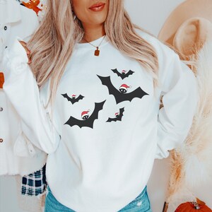 K&ouml;nnte beinhalten: Wei&szlig;er Pullover mit einem festlichen Design aus schwarzen Flederm&auml;usen, die Weihnachtsm&uuml;tzen tragen. Die Flederm&auml;use fliegen in verschiedene Richtungen.