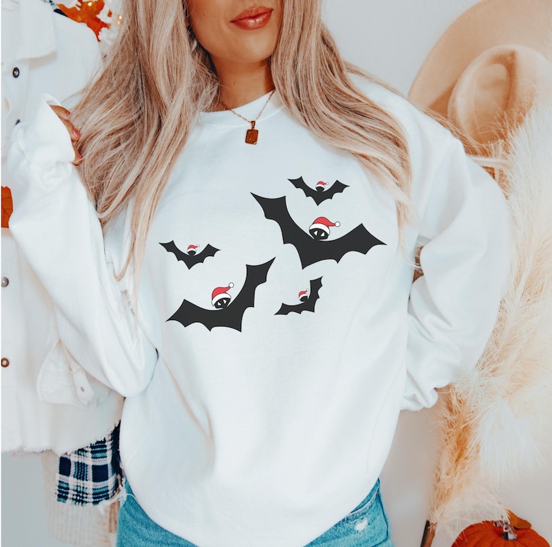 K&ouml;nnte beinhalten: Wei&szlig;er Pullover mit einem festlichen Design aus schwarzen Flederm&auml;usen, die Weihnachtsm&uuml;tzen tragen. Die Flederm&auml;use fliegen in verschiedene Richtungen.