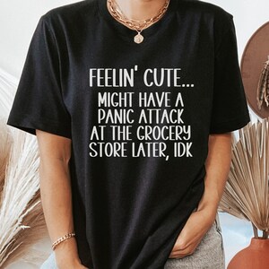 May include: Black t-shirt with white text that reads "Feelin' cute... Might have a panic attack at the grocery store later, idk."