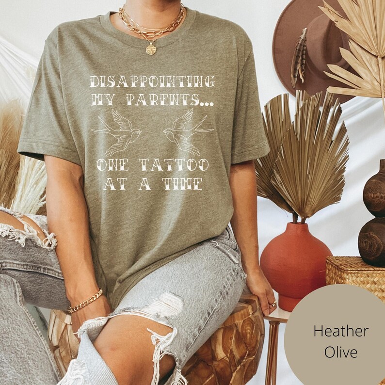 May include: A heather olive t-shirt with the text "Disappointing my parents... One tattoo at a time" in white lettering. Two stylized birds are above the text.