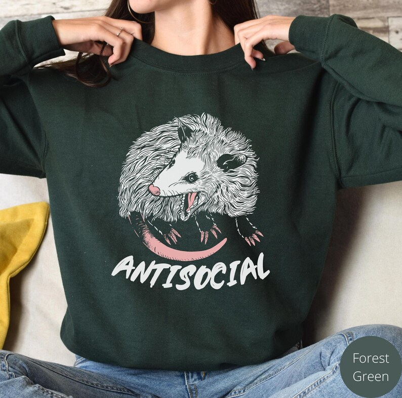 K&ouml;nnte beinhalten: Ein dunkelgr&uuml;nes Sweatshirt mit einer wei&szlig;en und schwarzen Illustration eines Opossums mit dem Text "ANTISOCIAL" in Rosa.
