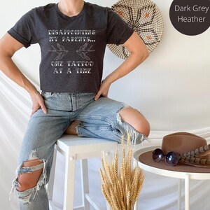 May include: A dark grey heather t-shirt with the text "DISAPPOINTING MY PARENTS... ONE TATTOO AT A TIME" and two stylized birds flying above the text.