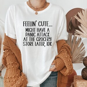 May include: White t-shirt with black text that reads "Feelin' Cute... Might Have A Panic Attack At The Grocery Store Later, IDK"