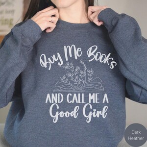 May include: Dark heather grey sweatshirt with the text "Buy Me Books And Call Me A Good Girl" in white. The text is surrounded by a stylized drawing of an open book with flowers.