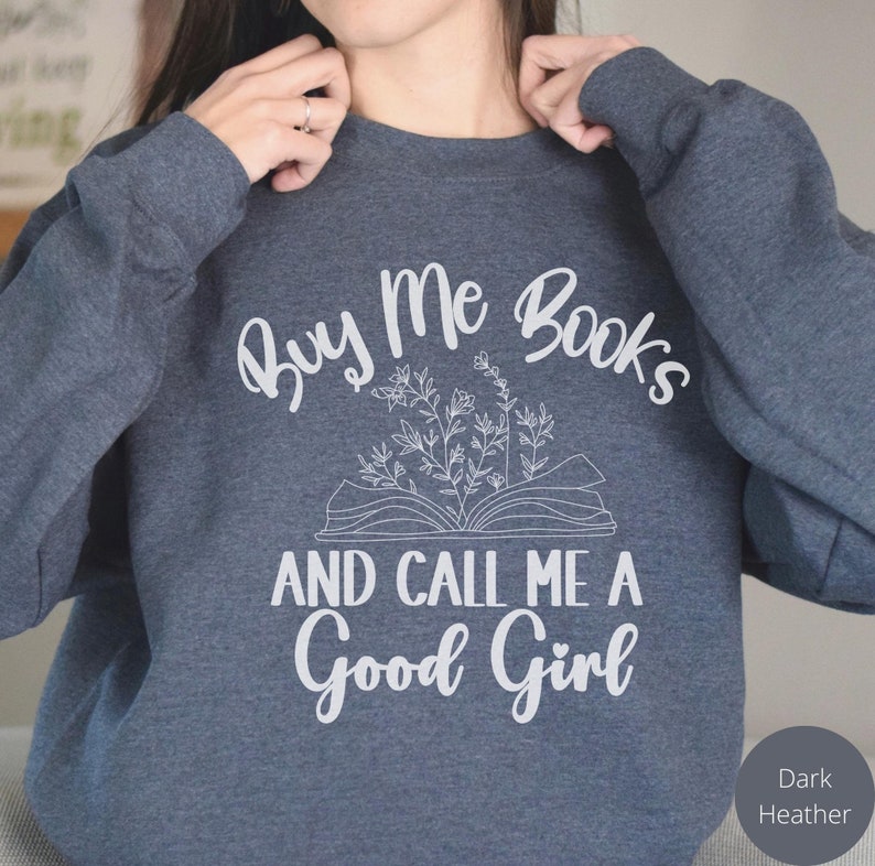 May include: Dark heather grey sweatshirt with the text "Buy Me Books And Call Me A Good Girl" in white. The text is surrounded by a stylized drawing of an open book with flowers.