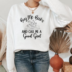 May include: White sweatshirt with the text "Buy Me Books and Call Me a Good Girl" in black lettering. The text is accompanied by a drawing of an open book with flowers.