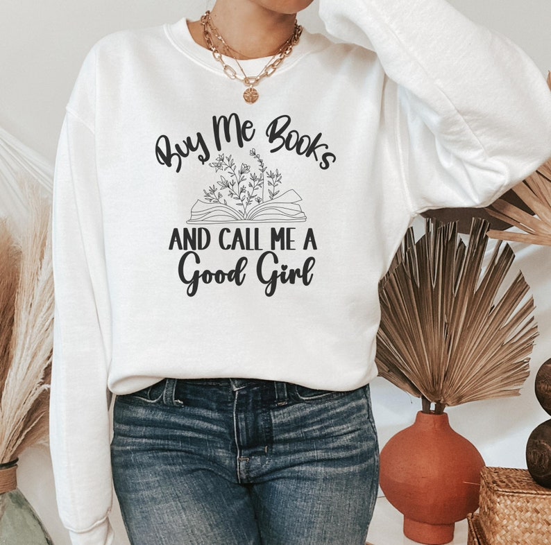 May include: White sweatshirt with the text "Buy Me Books and Call Me a Good Girl" in black lettering. The text is accompanied by a drawing of an open book with flowers.