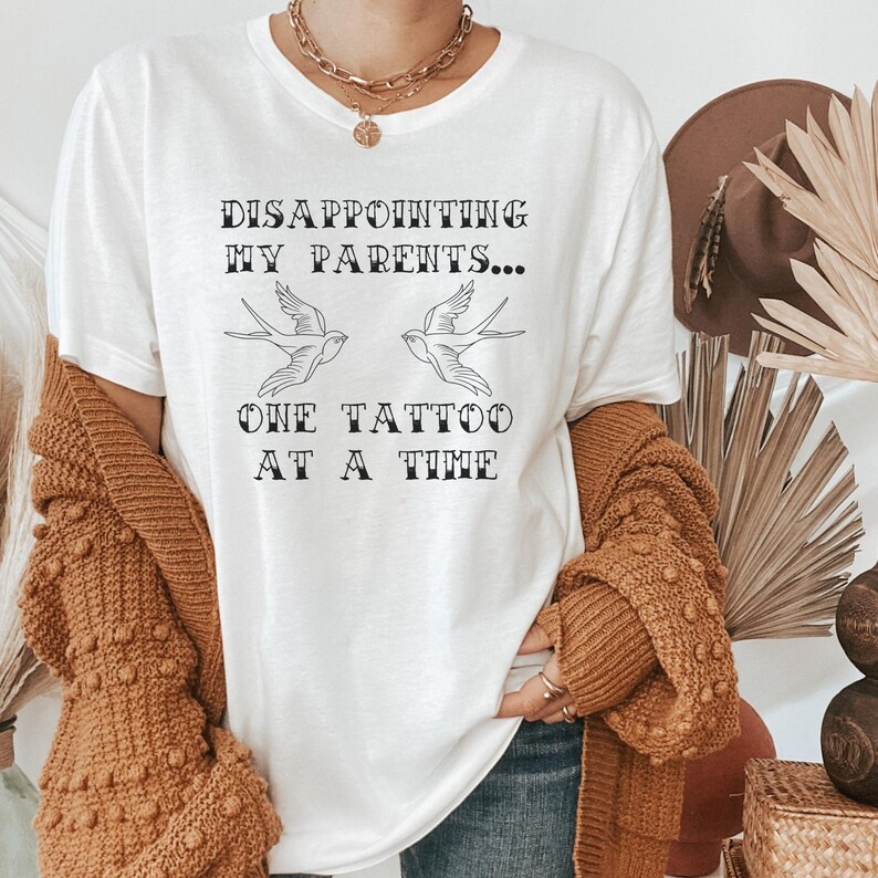 May include: White t-shirt with the text "DISAPPOINTING MY PARENTS... ONE TATTOO AT A TIME" and two stylized birds.