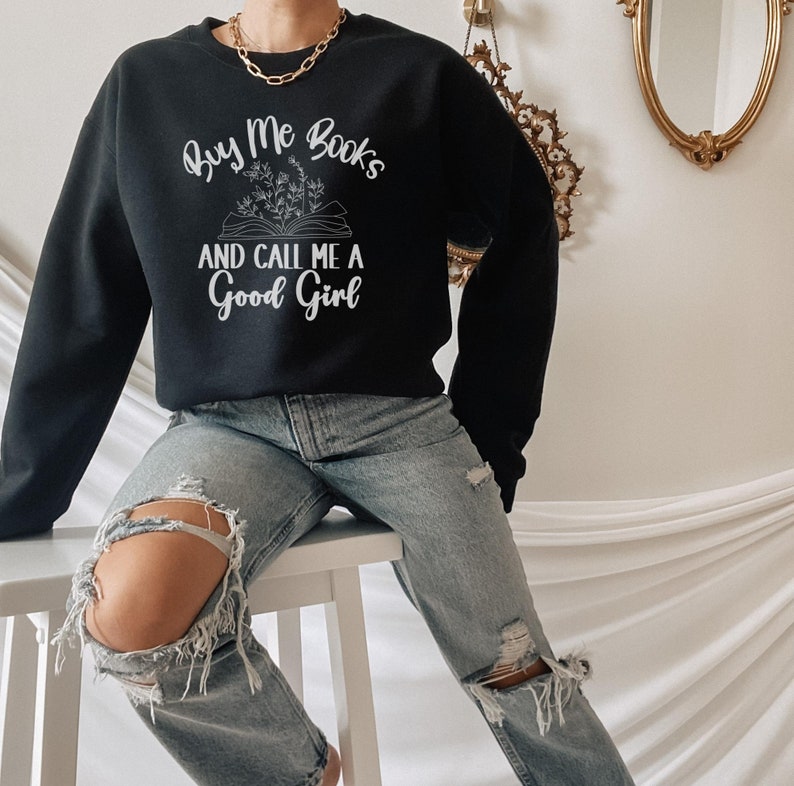 May include: A black sweatshirt with the text "Buy Me Books and Call Me a Good Girl" in white lettering. The text is accompanied by a drawing of a book with flowers on top.