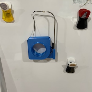 May include: A blue and white magnetic tape measure with a metal hook and a wire loop. The tape measure is attached to a white refrigerator with other magnets.