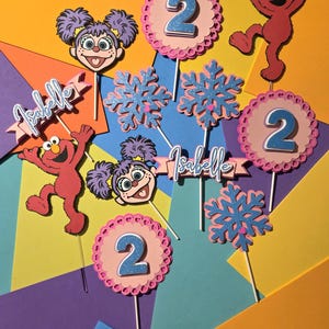 May include: Colorful birthday decorations featuring Elmo and a girl character, with the number 2. Includes snowflake and banner accents with the name "Isabelle". The decorations are on sticks, perfect for a cake or cupcakes.