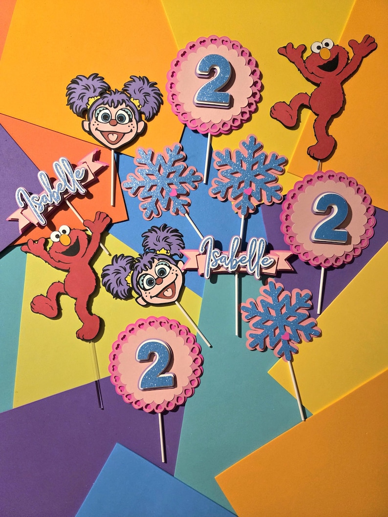 May include: Colorful birthday decorations featuring Elmo and a girl character, with the number 2. Includes snowflake and banner accents with the name "Isabelle". The decorations are on sticks, perfect for a cake or cupcakes.