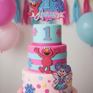 May include: A three-tiered birthday cake with pink, blue, and white frosting. The cake is decorated with Sesame Street characters, snowflakes, and the name "Amina" with the number "1". The cake sits on a pink cake stand.