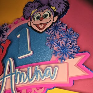 May include: A colorful birthday cake topper featuring a cartoon character with purple hair and a smiling face. The number "1" is displayed, along with the name "Amina" in a layered design. Decorative blue glitter snowflakes add a festive touch.