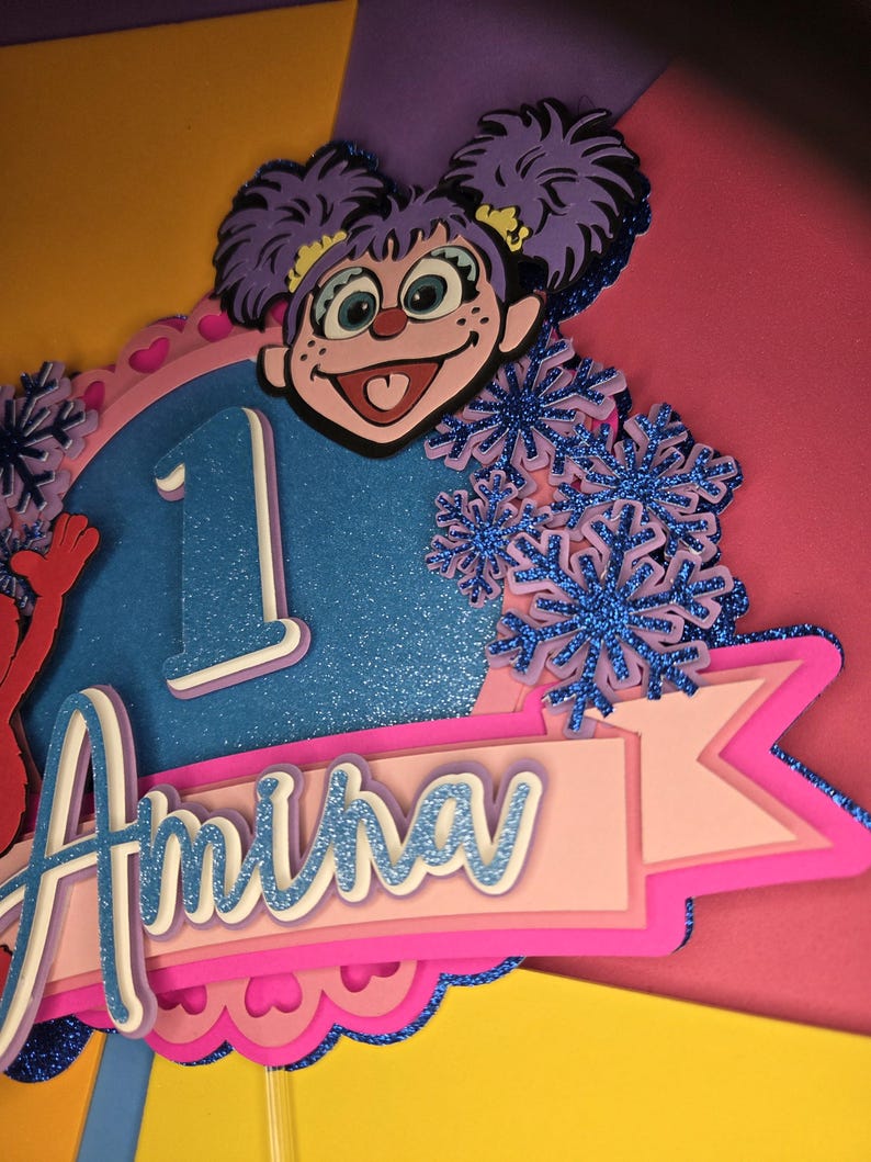 May include: A colorful birthday cake topper featuring a cartoon character with purple hair and a smiling face. The number "1" is displayed, along with the name "Amina" in a layered design. Decorative blue glitter snowflakes add a festive touch.