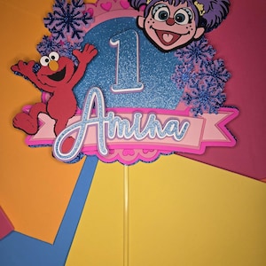 May include: A colorful cake topper featuring Elmo and a character with purple hair. The topper has a blue glitter background with the number 1 and the name "Amina" in a banner. Decorative snowflakes and hearts are also included.