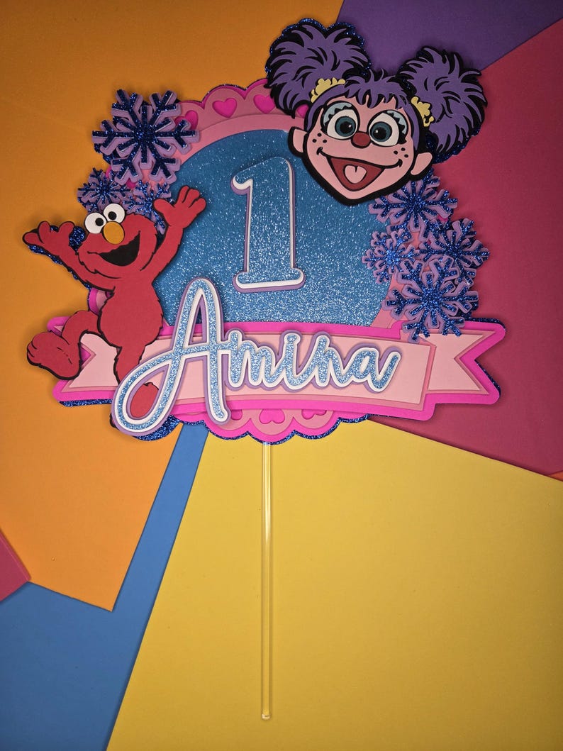 May include: A colorful cake topper featuring Elmo and a character with purple hair. The topper has a blue glitter background with the number 1 and the name "Amina" in a banner. Decorative snowflakes and hearts are also included.