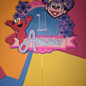 May include: A colorful cake topper featuring Elmo and a character with purple hair. The topper includes the number 1, the name "Amira", and snowflake accents. The design is set against a vibrant background.