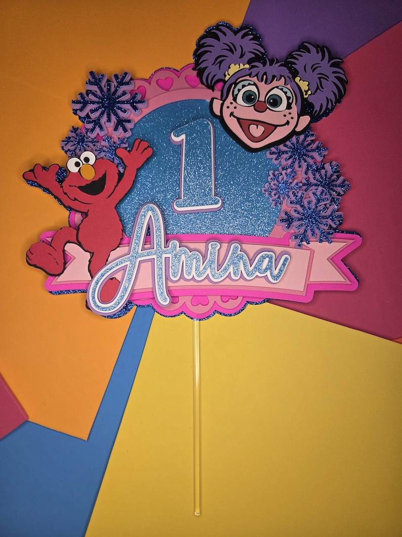 May include: A colorful cake topper featuring Elmo and a character with purple hair. The topper includes the number 1, the name "Amira", and snowflake accents. The design is set against a vibrant background.