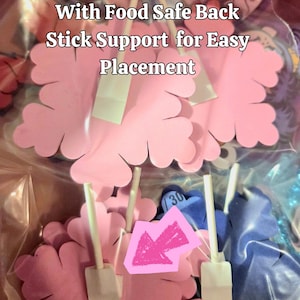 May include: Pink flower-shaped cake decorations with food-safe stick supports for easy placement. The image shows several pink flower decorations with white stick supports, and the text "Cake Decorations With Food Safe Back Stick Support for Easy Placement".